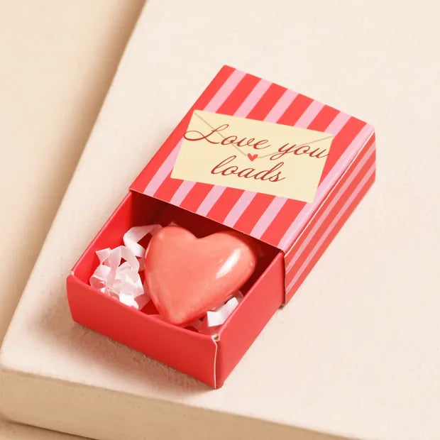 Pink ceramic heart inside open red-striped matchbox with envelope label on cream background
