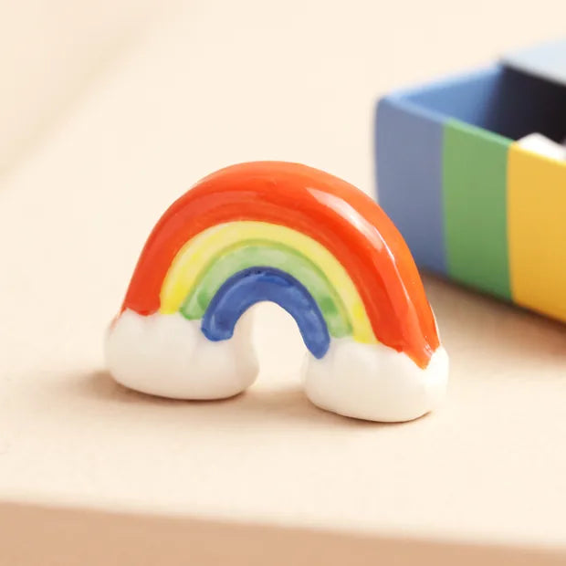 Ceramic rainbow token with colourful arcs and white clouds beside striped matchbox on cream background