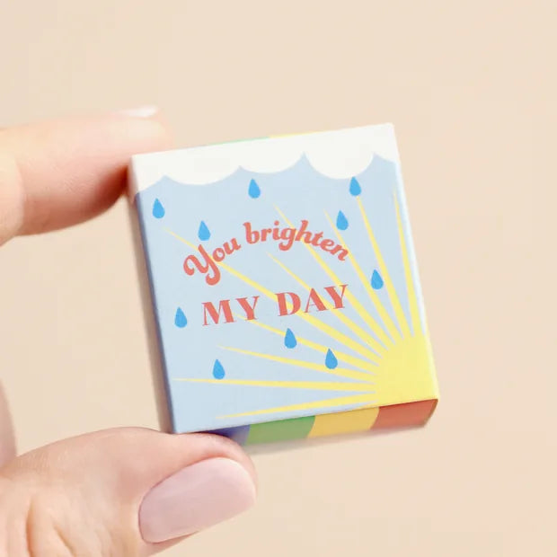 Hand holding closed rainbow matchbox with sunshine design on cream background