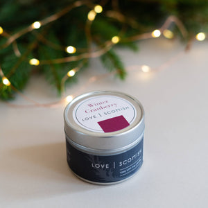 Winter Cranberry travel tin candle by Love Scottish on a light surface with festive bokeh lights and pine branches.