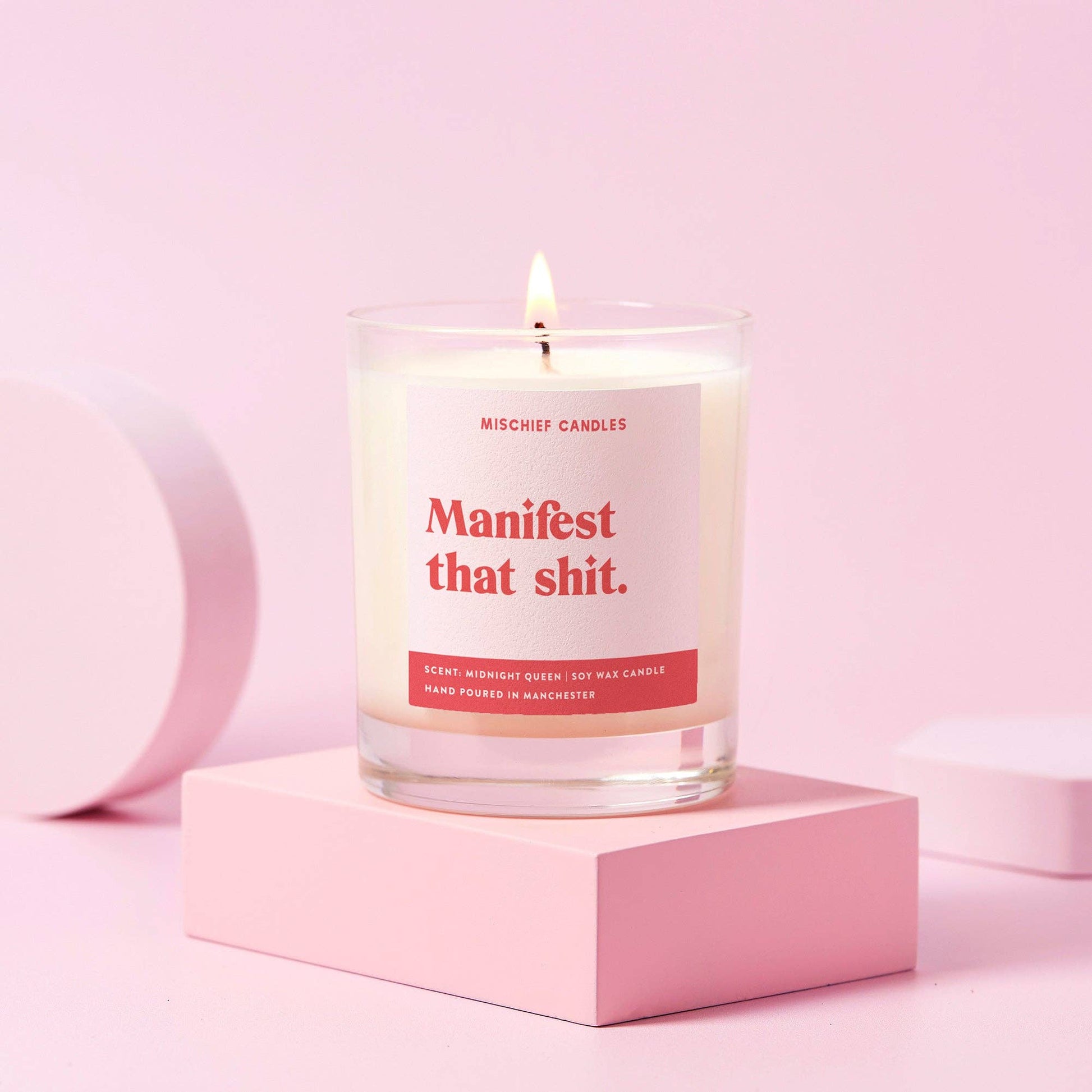 Manifest positivity candle in clear glass jar burning on a pink block backdrop