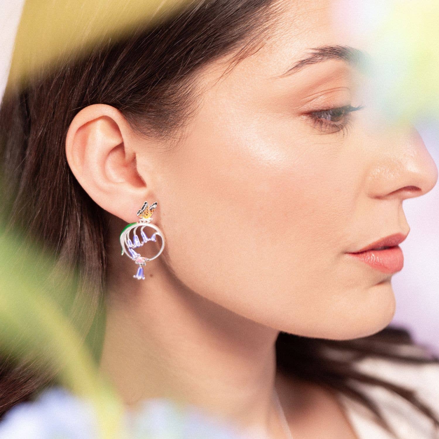 Model wearing Bluebell & Butterfly Hoop Earrings with blue floral background, highlighting delicate enamel detail.