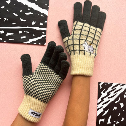 Hands wearing Moomin Chequered Gloves in green and cream knit with embroidered Moomin family design, against pink background.
