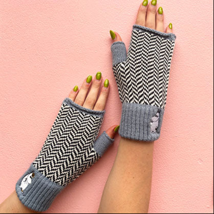 Hands wearing Moomin Herringbone Gloves in grey and black knit with embroidered Moomin detail, against pink background.

