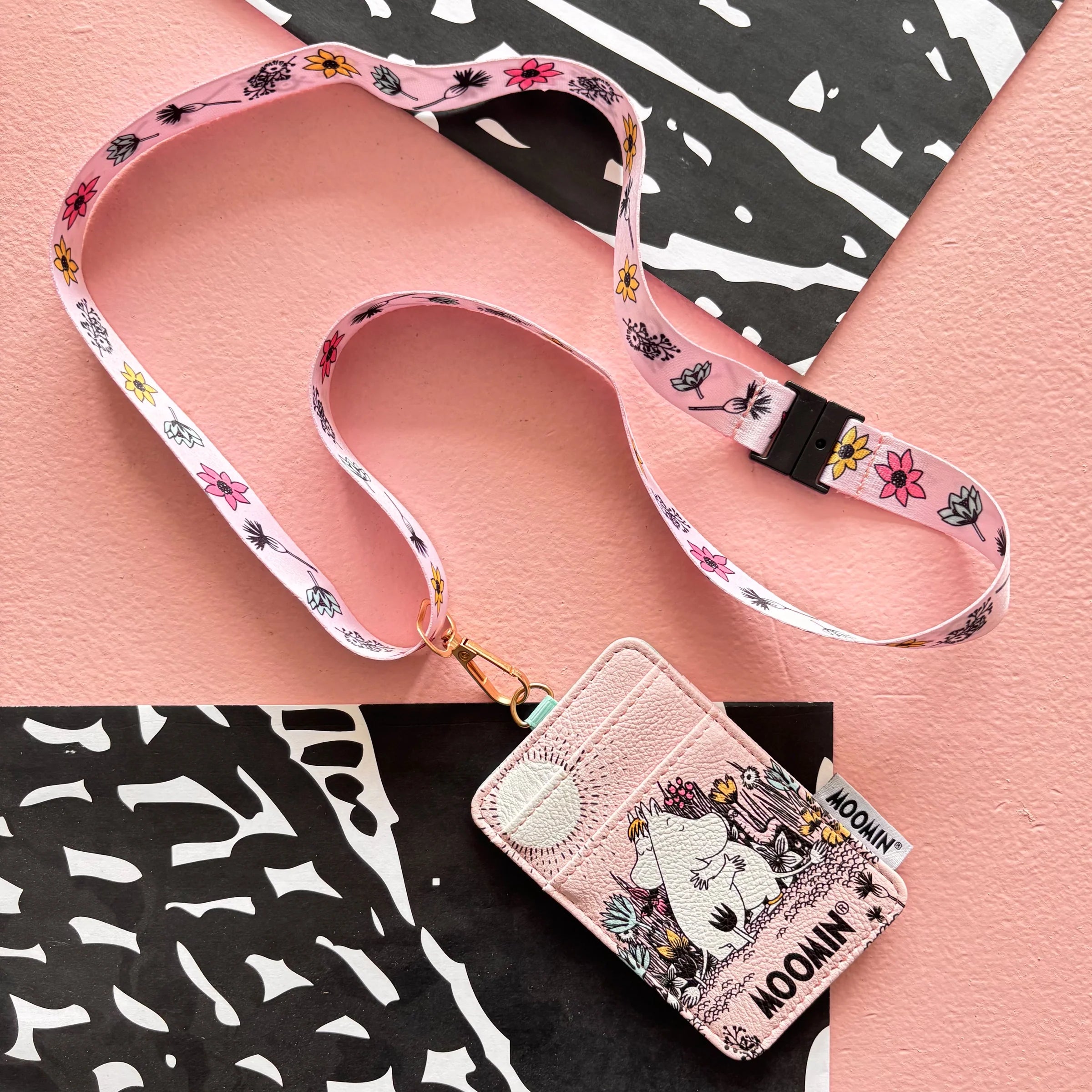 Flat lay of Moomin Love Lanyard with floral print strap and cardholder on pink surface.
