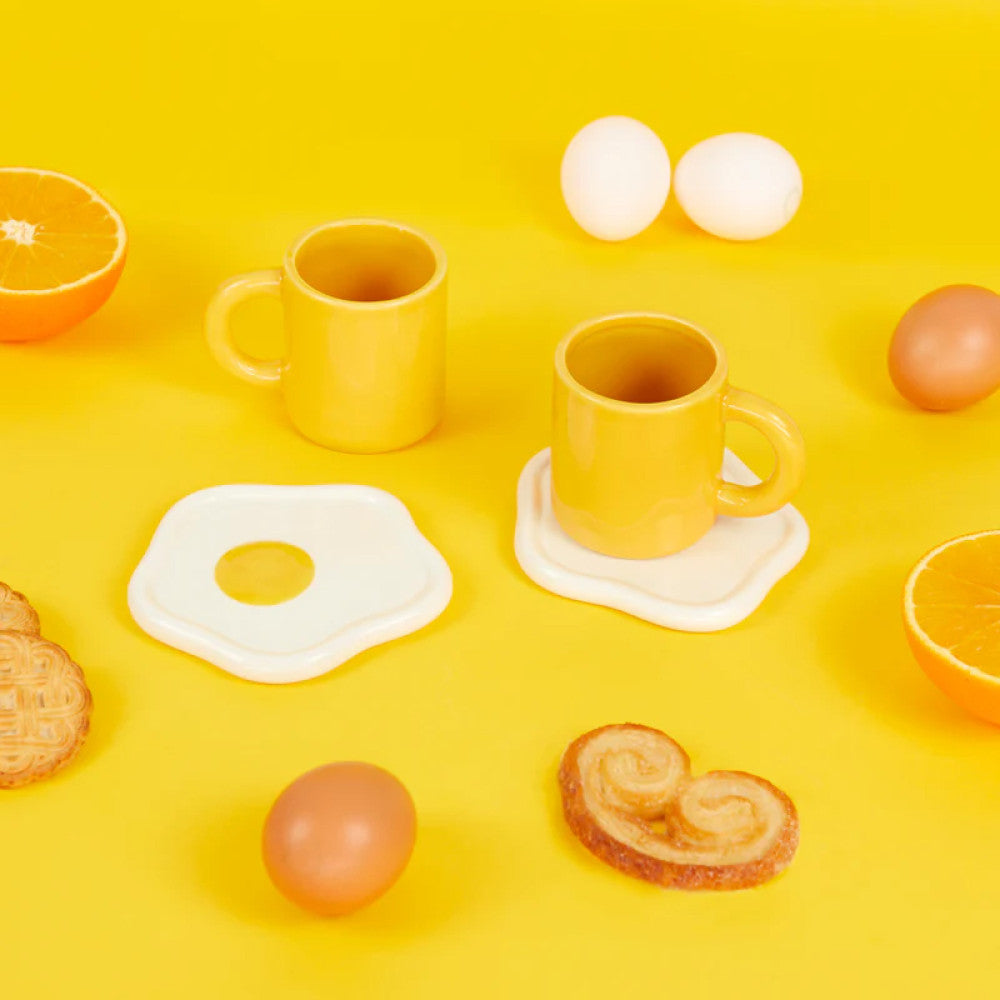 Yellow cups with fried egg saucers on yellow background with fruit
