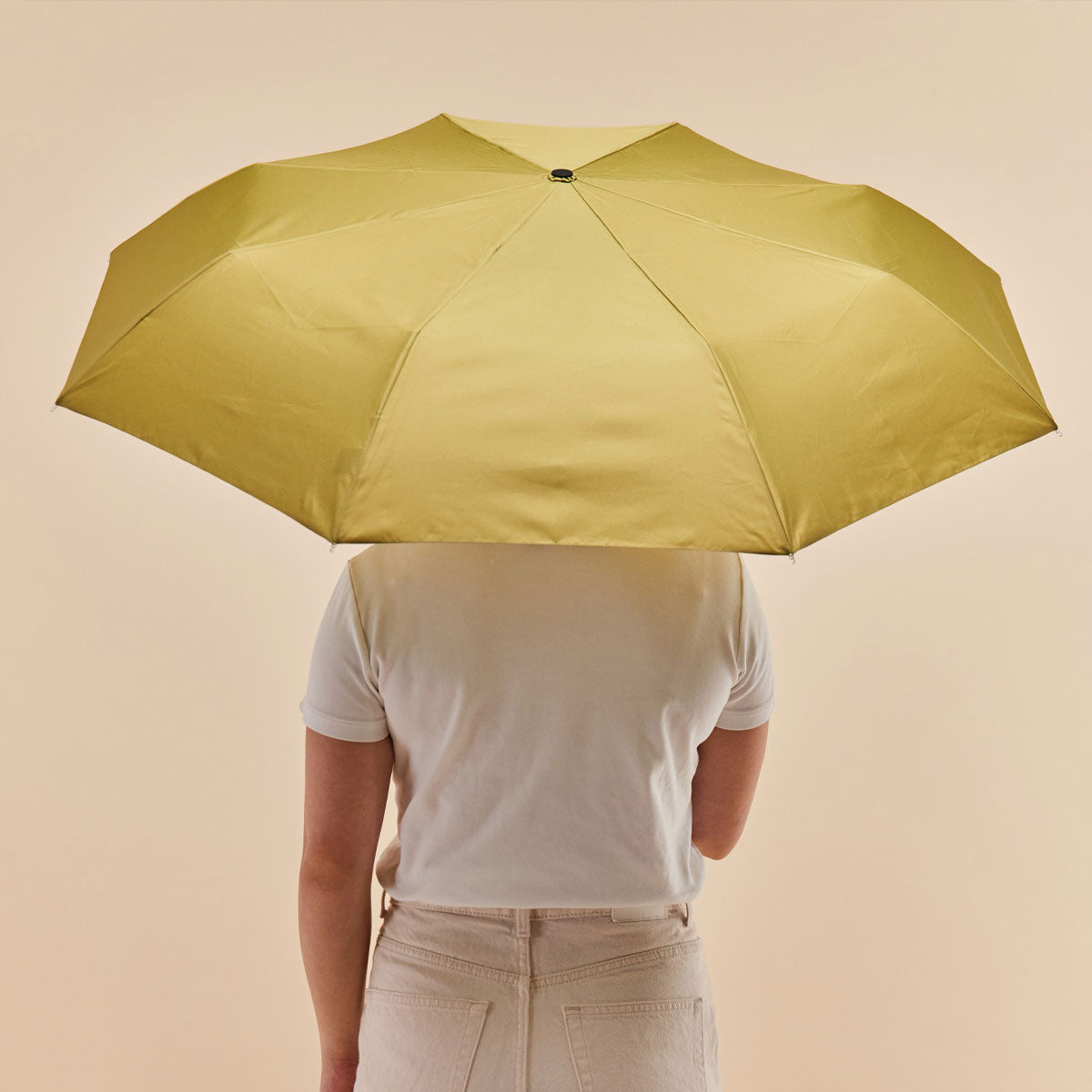 Olive duck umbrella open held from behind by model on warm beige background
