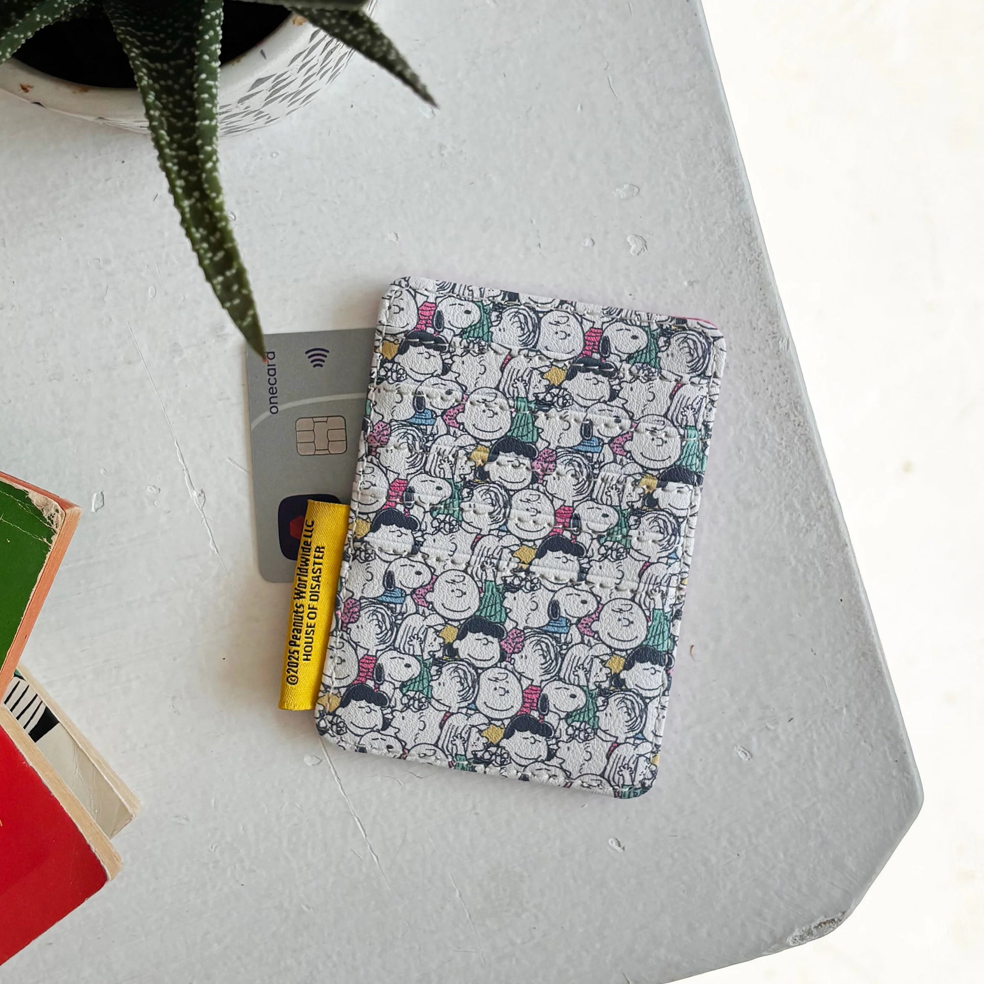 Back of Peanuts Lucy Cardholder with multicoloured Peanuts character print beside green plant.