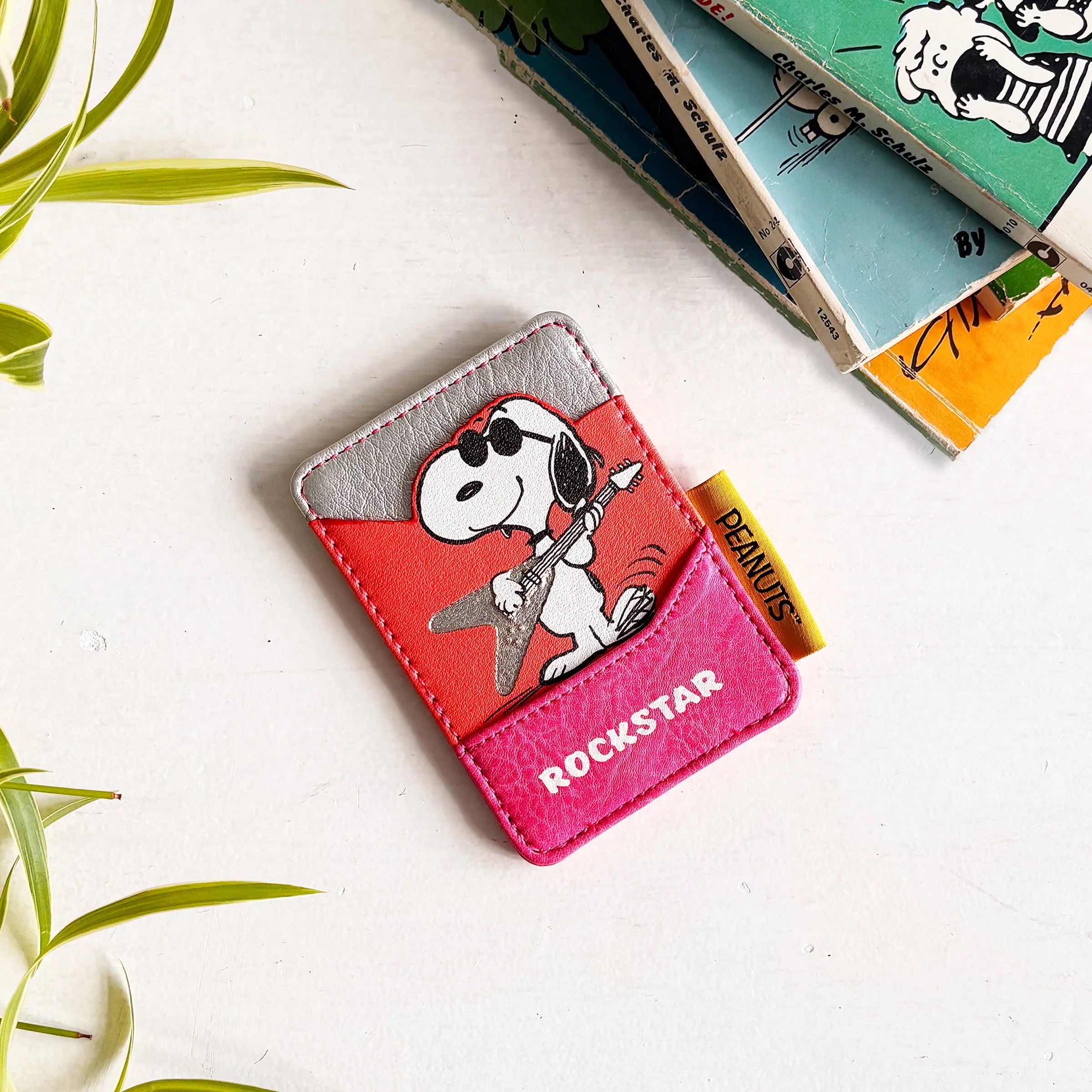 Peanuts Rockstar Cardholder featuring Snoopy playing guitar in red, pink, and silver design beside Peanuts comic books.