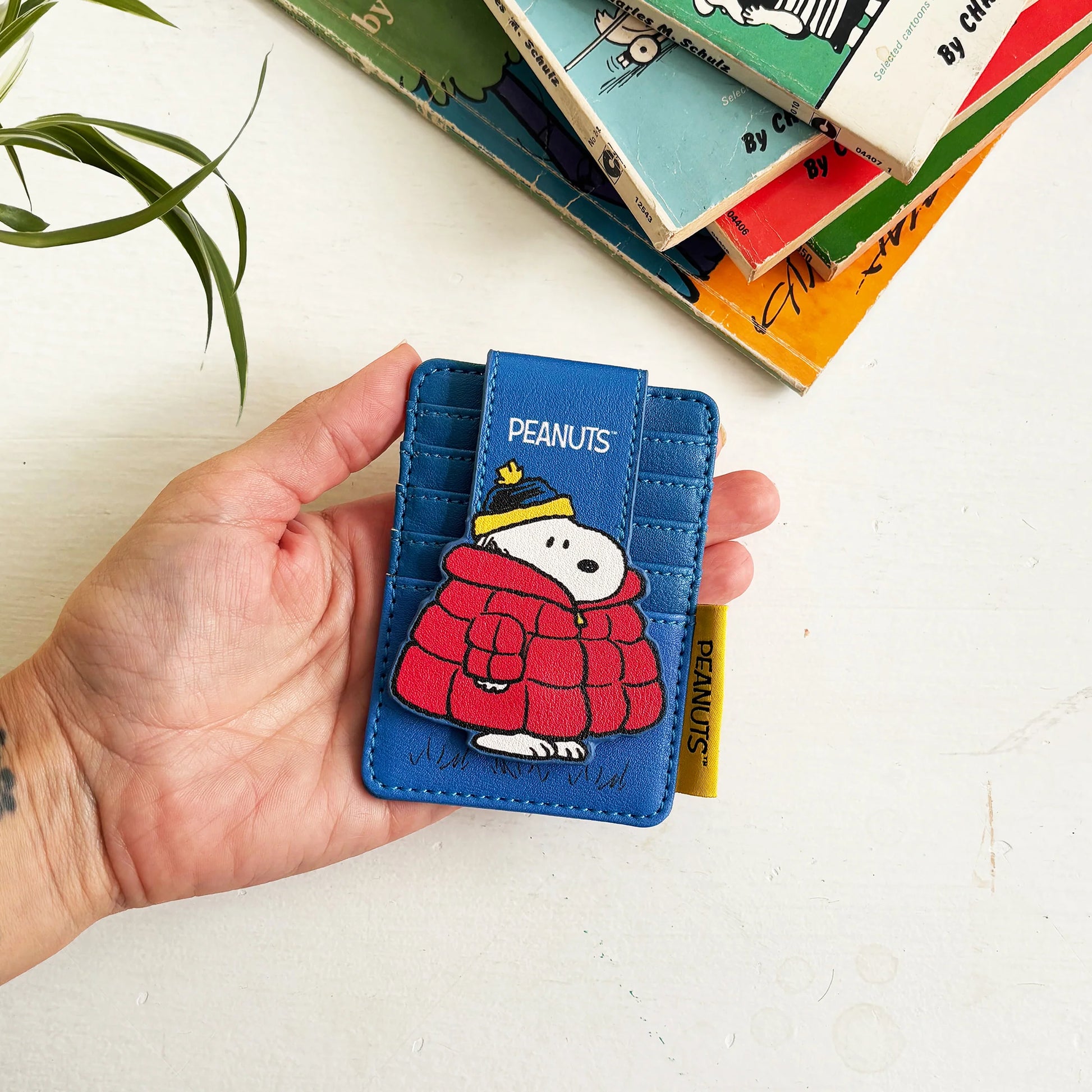 Hand holding Peanuts Snoopy Coat Foldover Card Holder featuring Snoopy in red puffer jacket on blue background.