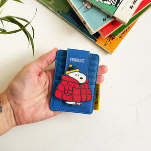 Hand holding Peanuts Snoopy Coat Foldover Card Holder featuring Snoopy in red puffer jacket on blue background.