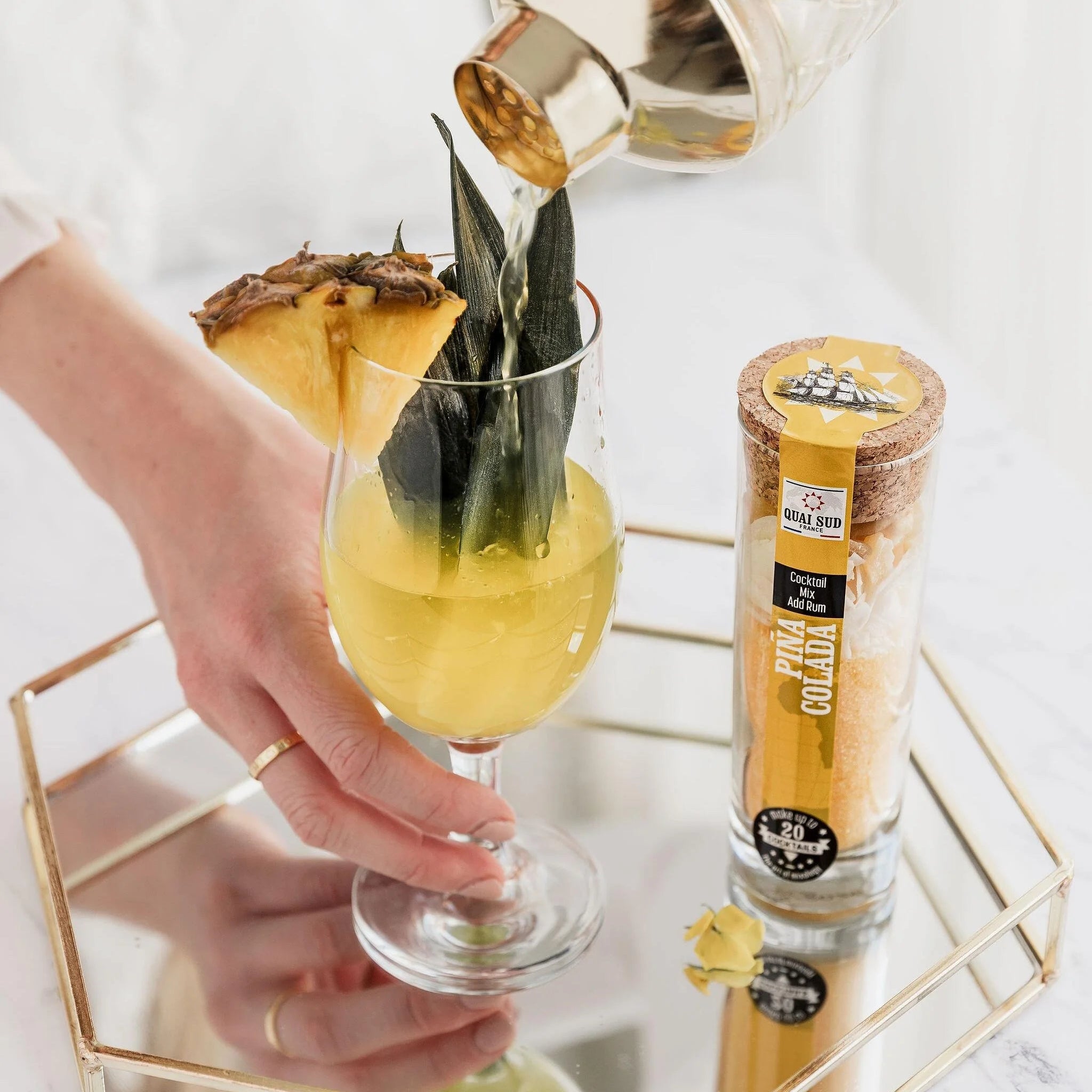 Pouring a freshly shaken Piña Colada cocktail over ice with pineapple garnish beside the cocktail mix tube.
