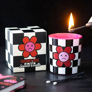 Sad Flower Coconut Rose Candle with checkerboard design and pink flower face, lit beside matching gift box on black background.