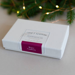 Winter Cranberry wax melts gift box labelled “Love Scottish” on a neutral surface with festive pine branches and warm fairy lights in the background.