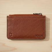 Brown leather zip-top card holder back view with Yoshi branding on wood surface