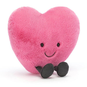 The image shows a bright pink heart-shaped plush toy with black embroidered eyes and a smile. The plushie is made of soft and fuzzy material and has a cuddly texture. The toy sits upright, with legs in front.