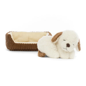 Cute and cuddly Jellycat Napping Nipper Dog, a sandy-brown plush dog with floppy ears, snuggled up in a cozy cordy brown bed.