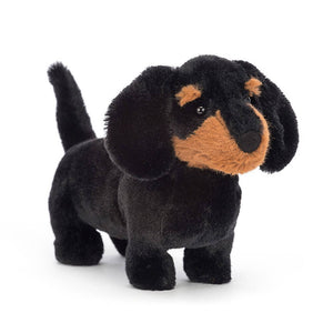 Jellycat Freddie Sausage Dog small children’s soft toy. He has a black furry body but a ginger and black patchwork face.