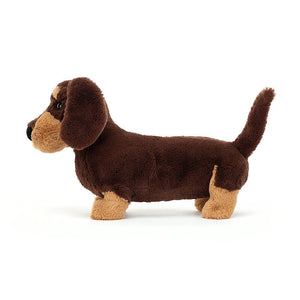 Large Jellycat Ottos Sausage Dog children’s soft toy. He is covered in dark brown fur and has caramel patches on his face and paws. 